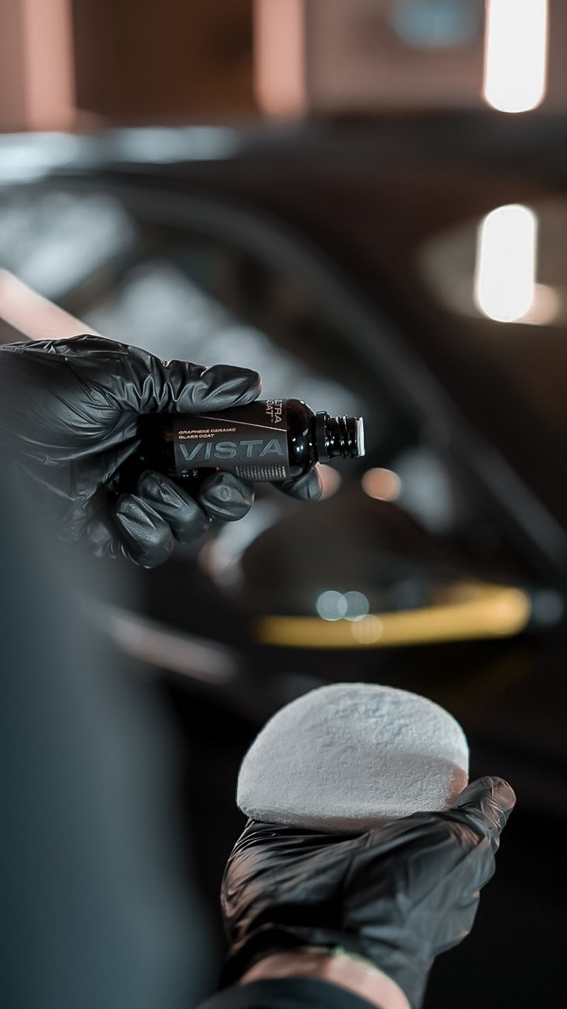 ULTRACOAT Vista InteliGraph graphene-ceramic glass coat held by a gloved hand, applicator pad ready, dark studio backdrop